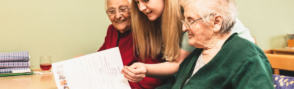 Eine Mitarbeiterin blättert mit zwei Bewohnerinnen durch eine Zeitschrift